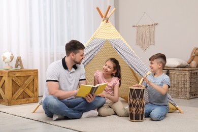 Photo of Father reading book to children near toy wigwam at home