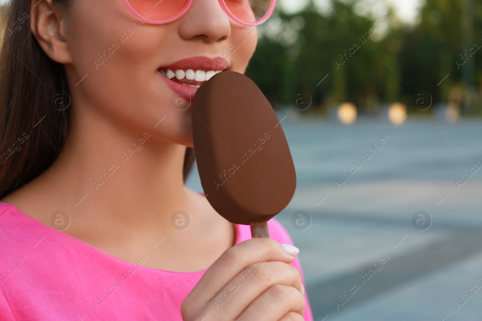 Photo of Young woman eating ice cream glazed in chocolate on city street, closeup