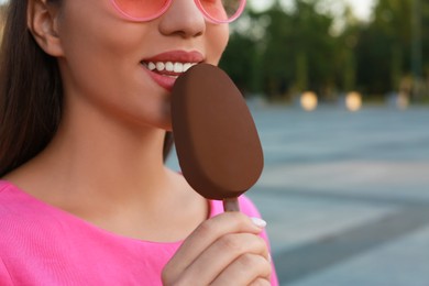 Photo of Young woman eating ice cream glazed in chocolate on city street, closeup
