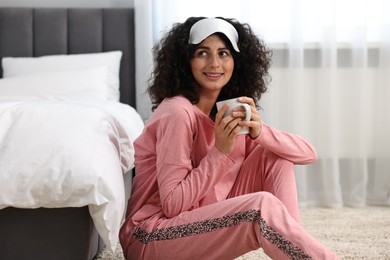 Photo of Beautiful young woman in stylish pyjama and sleep mask with cup of drink on floor at home