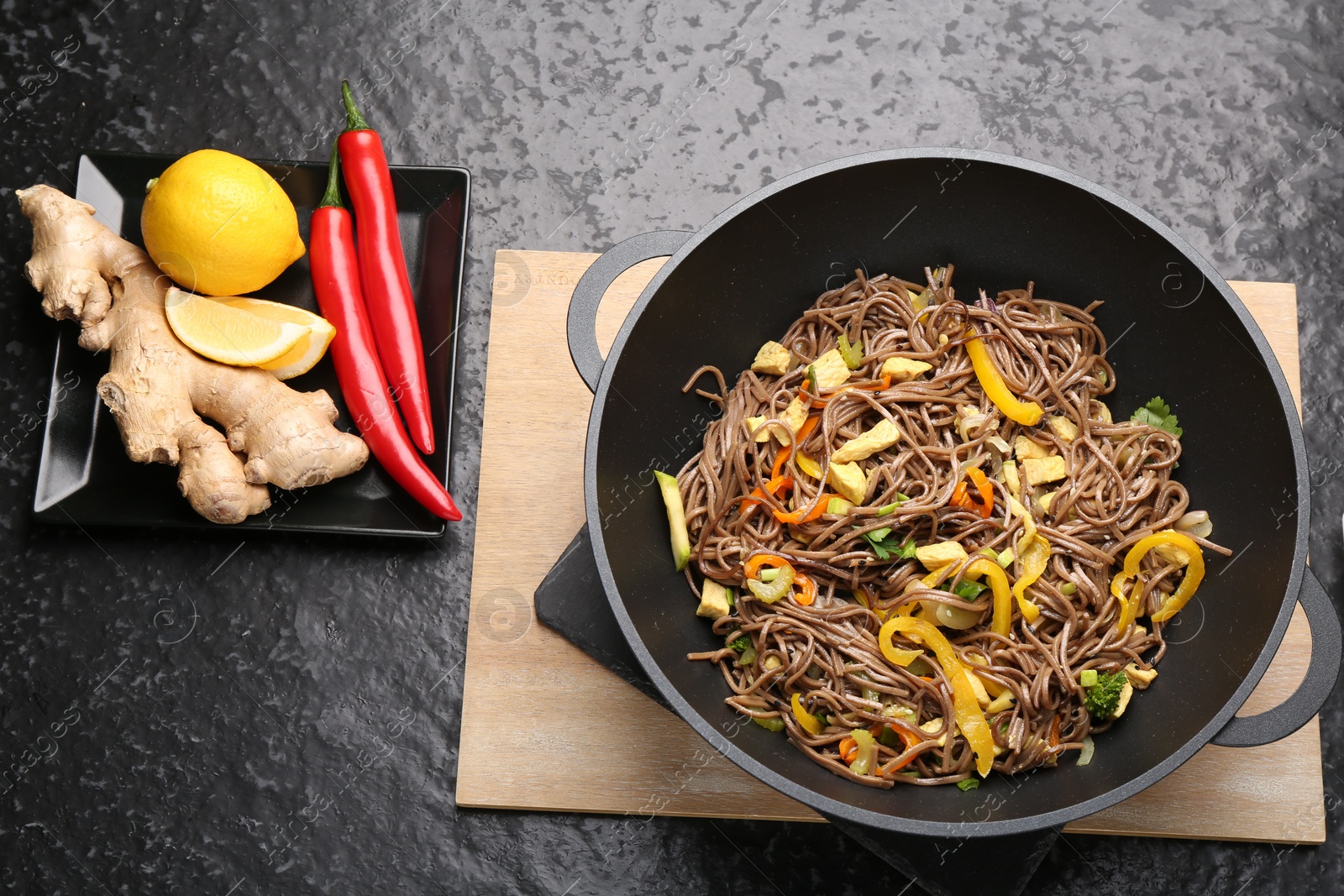 Photo of Stir-fry. Tasty noodles with vegetables, meat in wok and ingredients on dark textured table, flat lay