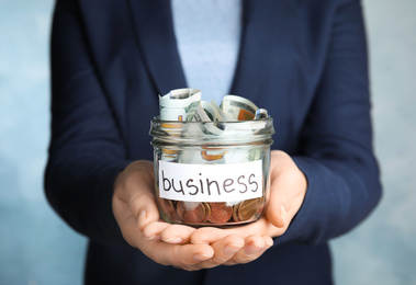Photo of Woman holding glass jar with money and tag BUSINESS on light blue background, closeup