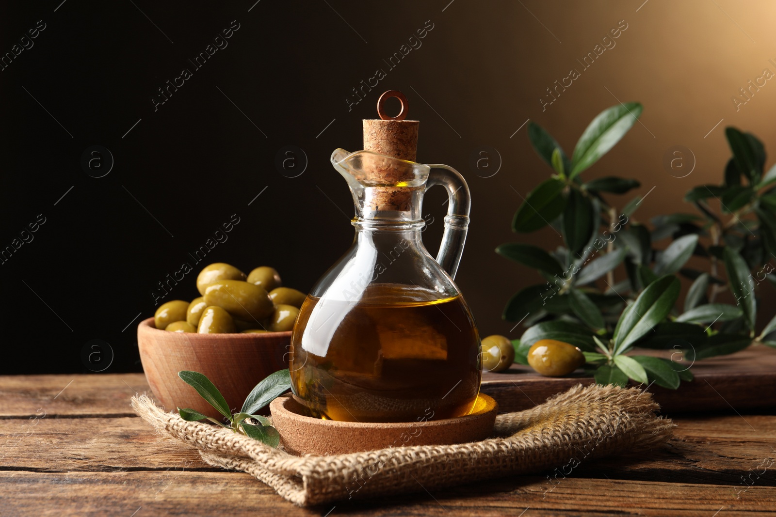 Photo of Glass jug of oil, ripe olives and green leaves on wooden table