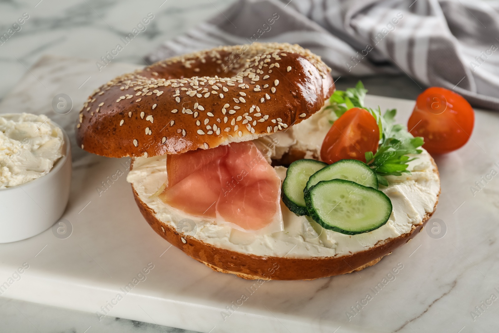 Photo of Delicious bagel with cream cheese, jamon, cucumber, tomato and parsley on white board, closeup