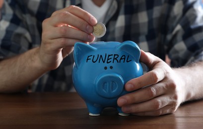 Image of Money for funeral expenses. Man putting coin into piggy bank at wooden table, closeup