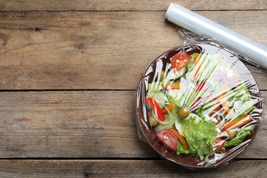 Photo of Bowl of fresh salad with plastic food wrap on wooden table, flat lay. Space for text