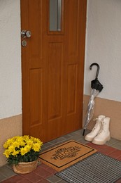 Door mat with word Welcome, boots, umbrella and beautiful flowers near entrance