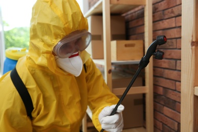Photo of Pest control worker in protective suit spraying pesticide indoors
