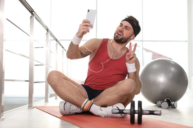Photo of Lazy young man with sport equipment taking selfie indoors