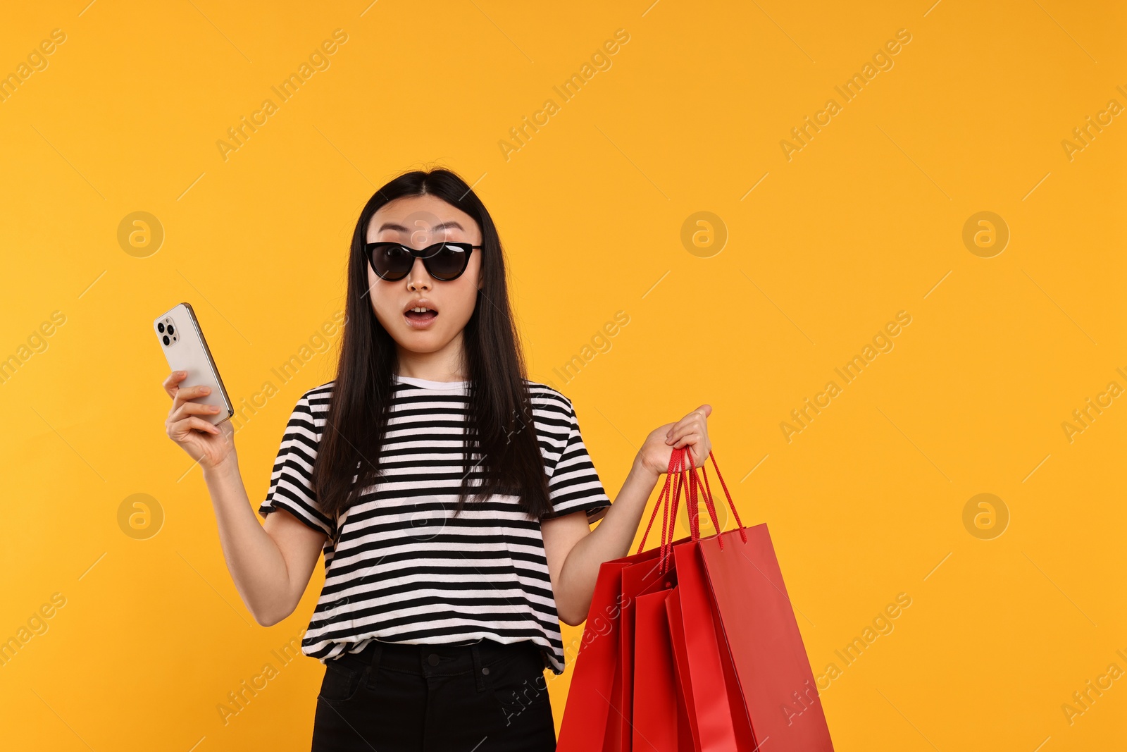 Photo of Surprised woman with shopping bags and smartphone on yellow background. Space for text