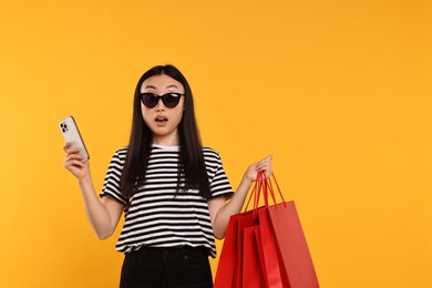 Surprised woman with shopping bags and smartphone on yellow background. Space for text