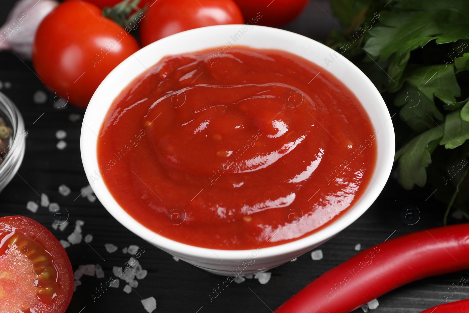 Photo of Delicious ketchup in bowl, products and salt on black wooden table, closeup. Tomato sauce