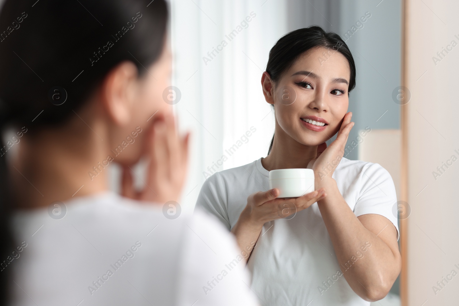 Photo of Happy woman applying face cream near mirror at home