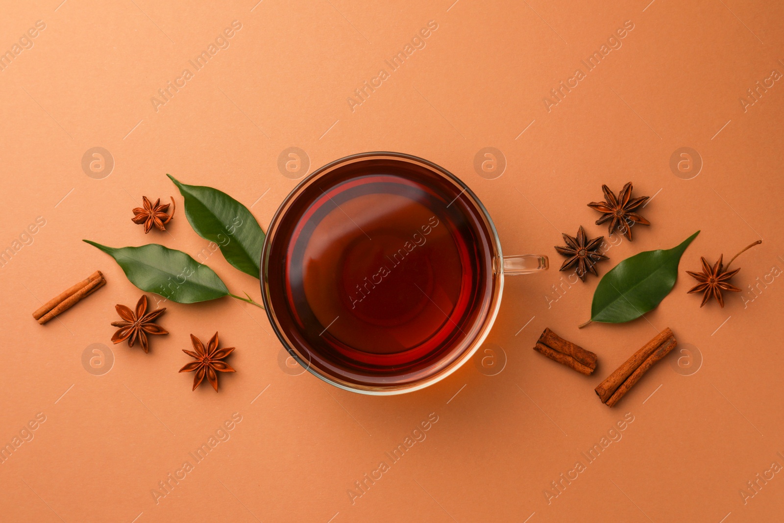 Photo of Cup of tea, anise stars, green leaves and cinnamon sticks on brown background, flat lay