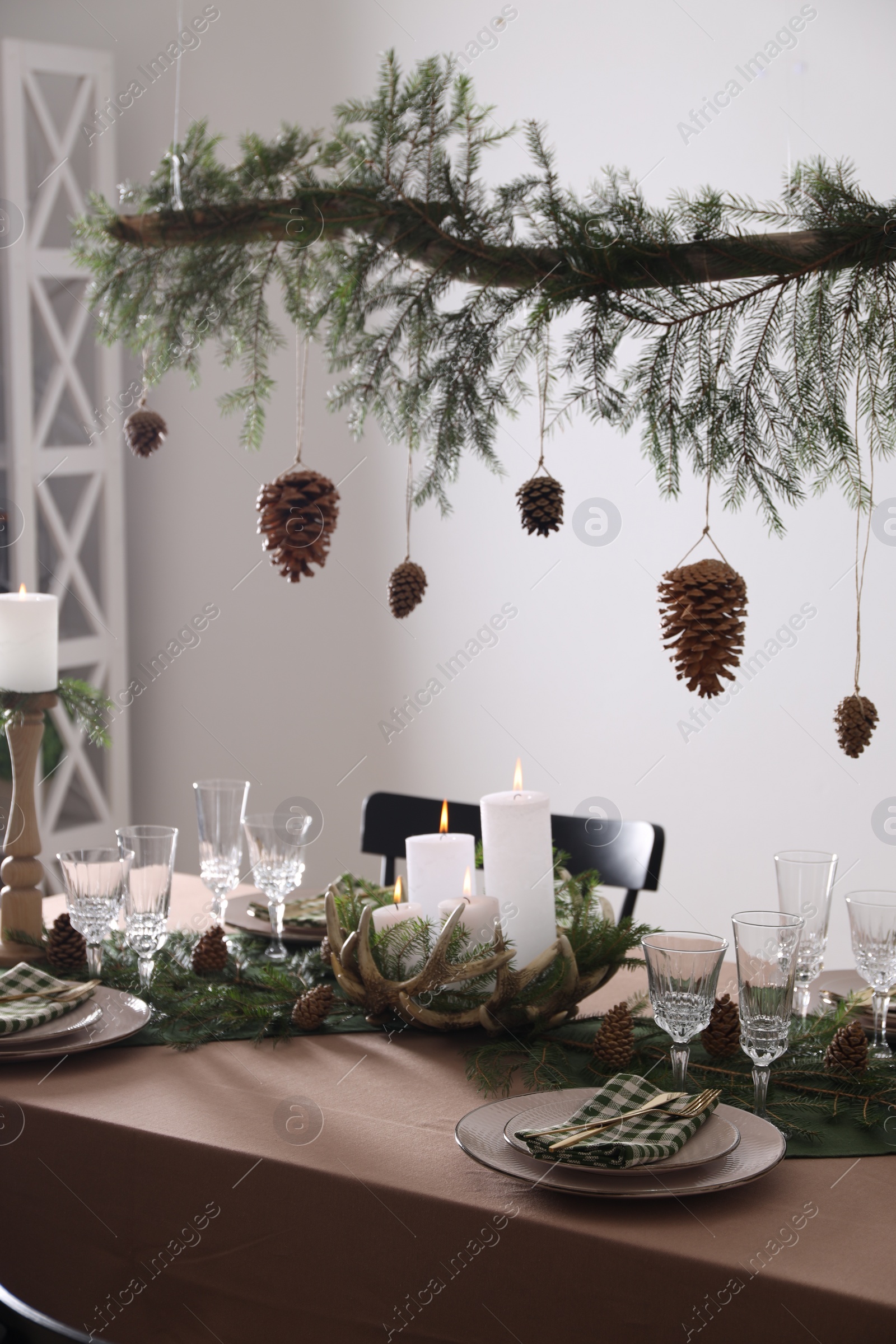 Photo of Christmas celebration. Cones hanging from fir tree branch over table with burning candles and tableware