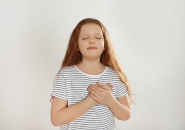 Cute grateful little girl with hands on chest against light background