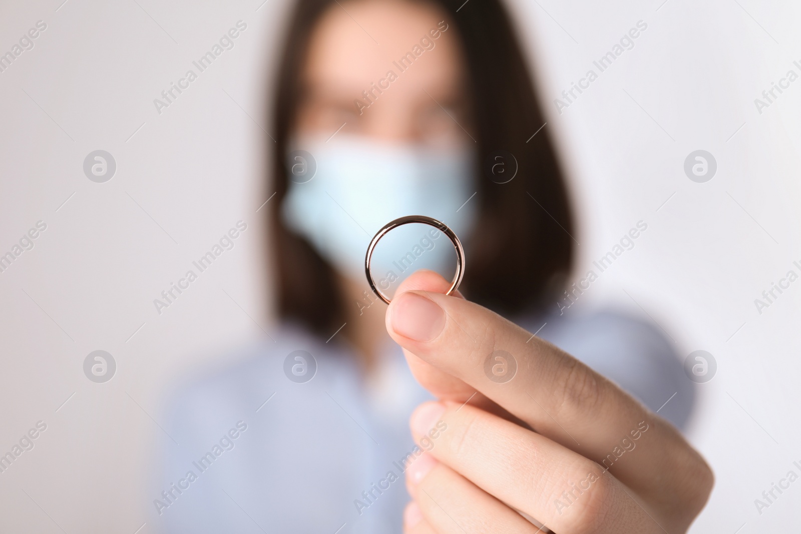 Photo of Woman in protective mask holding wedding ring against light background, focus on hand. Divorce during coronavirus quarantine