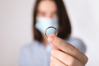 Photo of Woman in protective mask holding wedding ring against light background, focus on hand. Divorce during coronavirus quarantine