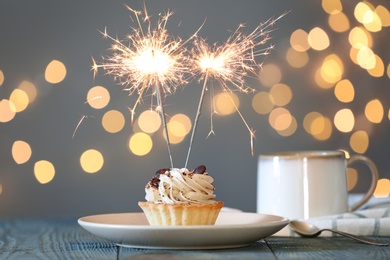 Photo of Cupcake with burning sparkler on table against blurred festive lights
