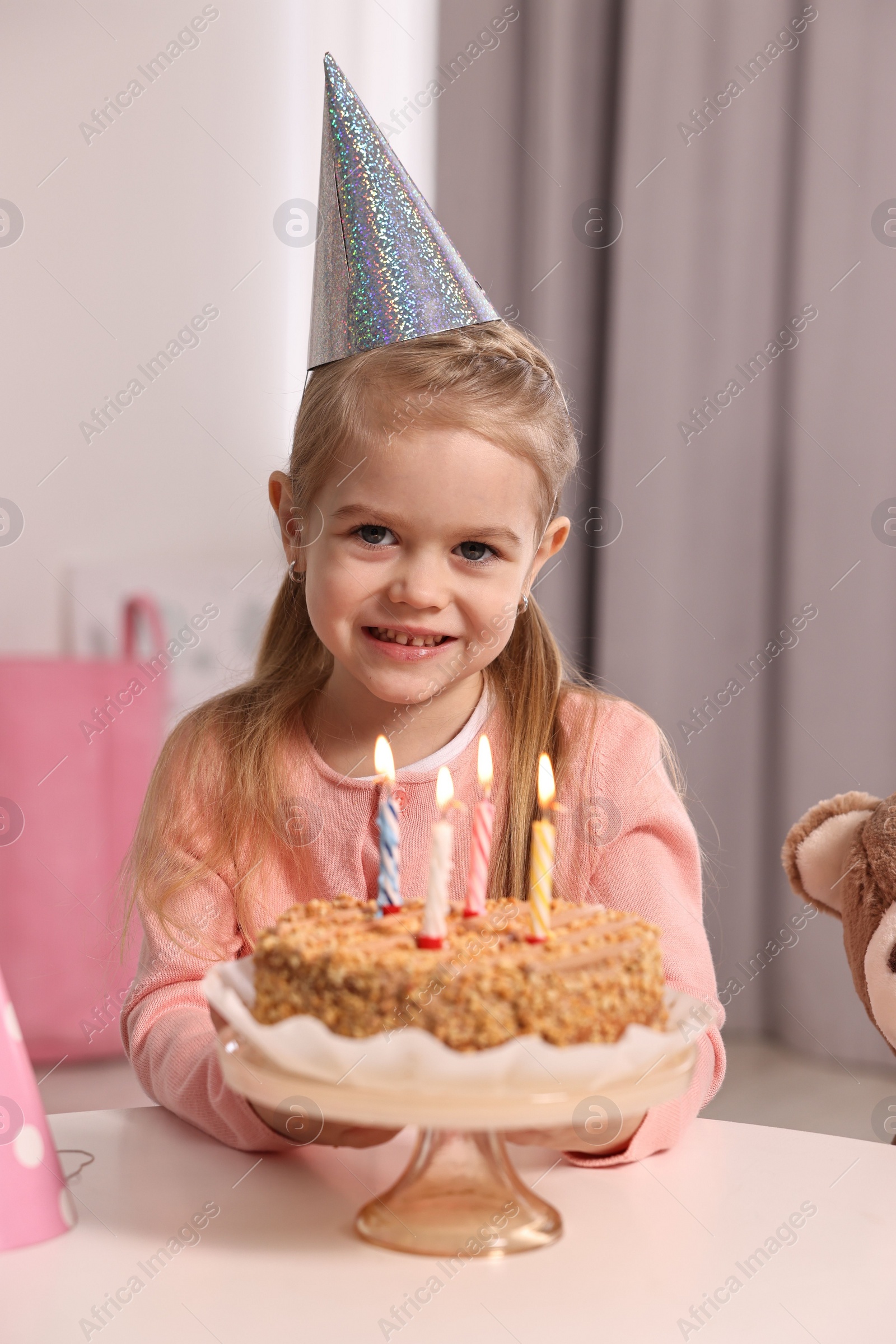 Photo of Cute girl in party hat with birthday cake at table indoors