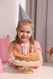 Cute girl in party hat with birthday cake at table indoors
