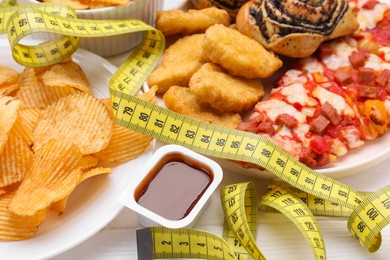 Different unhealthy food and measuring tape on white wooden table, closeup. Weight loss concept
