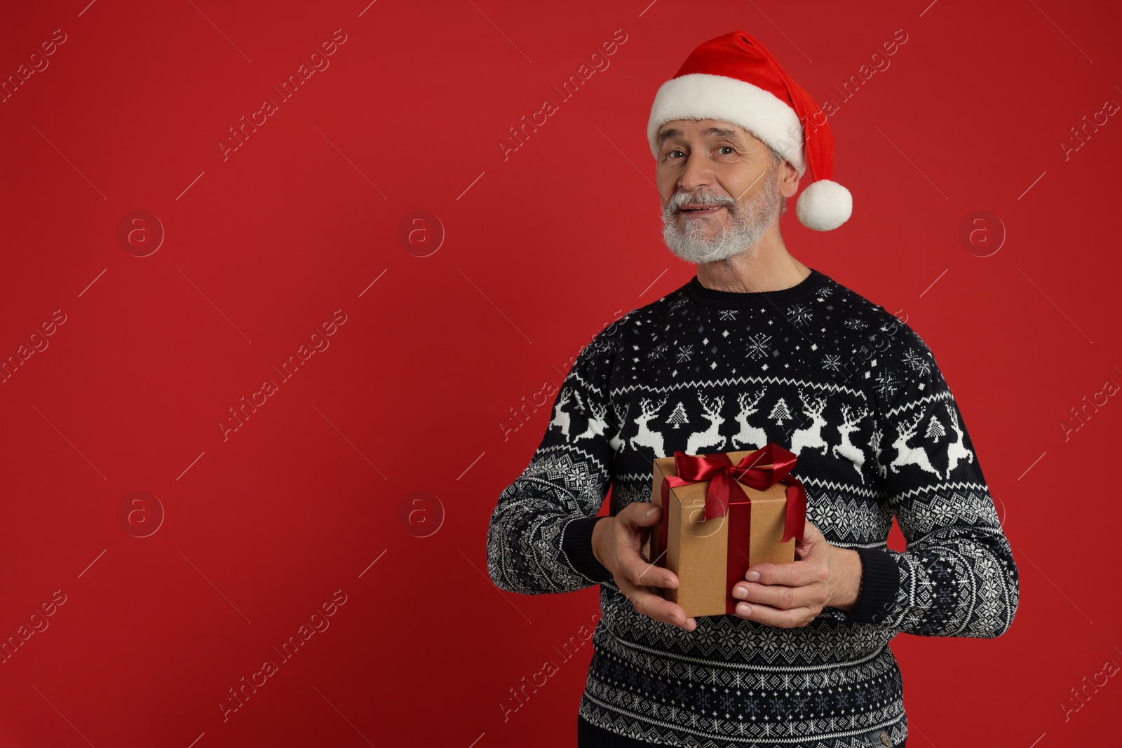 Photo of Senior man in Christmas sweater and Santa hat holding gift on red background. Space for text
