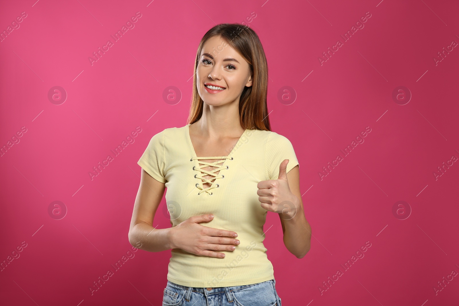 Photo of Healthy woman holding hand on belly and showing thumbs up gesture against pink background