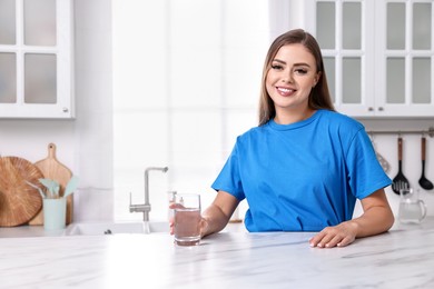 Photo of Happy woman with glass of fresh water at white marble table in kitchen, space for text