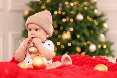 Photo of Cute little baby with Christmas bauble on red blanket against blurred festive lights, space for text. Winter holiday