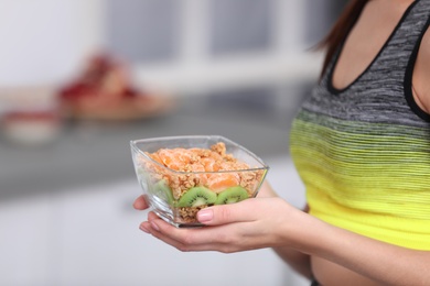 Photo of Young woman in fitness clothes holding bowl of cereal breakfast with fruits at home, closeup