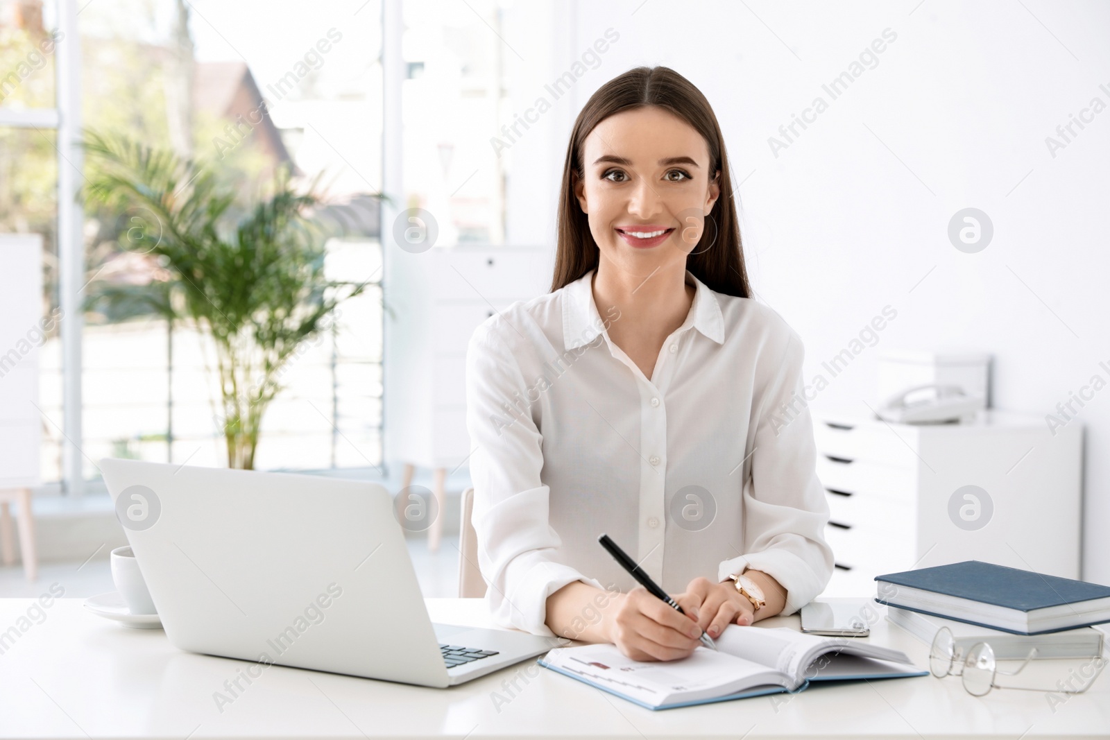 Photo of Young businesswoman writing in notebook while using laptop at table in office