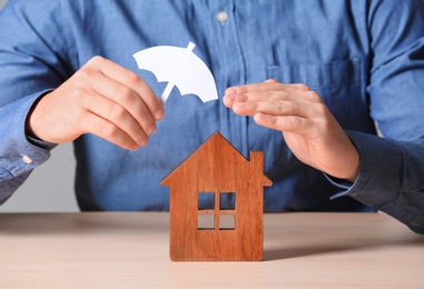 Photo of Man covering wooden house with umbrella cutout at table, closeup. Home insurance