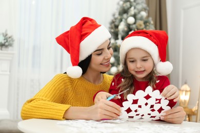 Photo of Mother and daughter in Santa hats making paper snowflake near Christmas tree at home