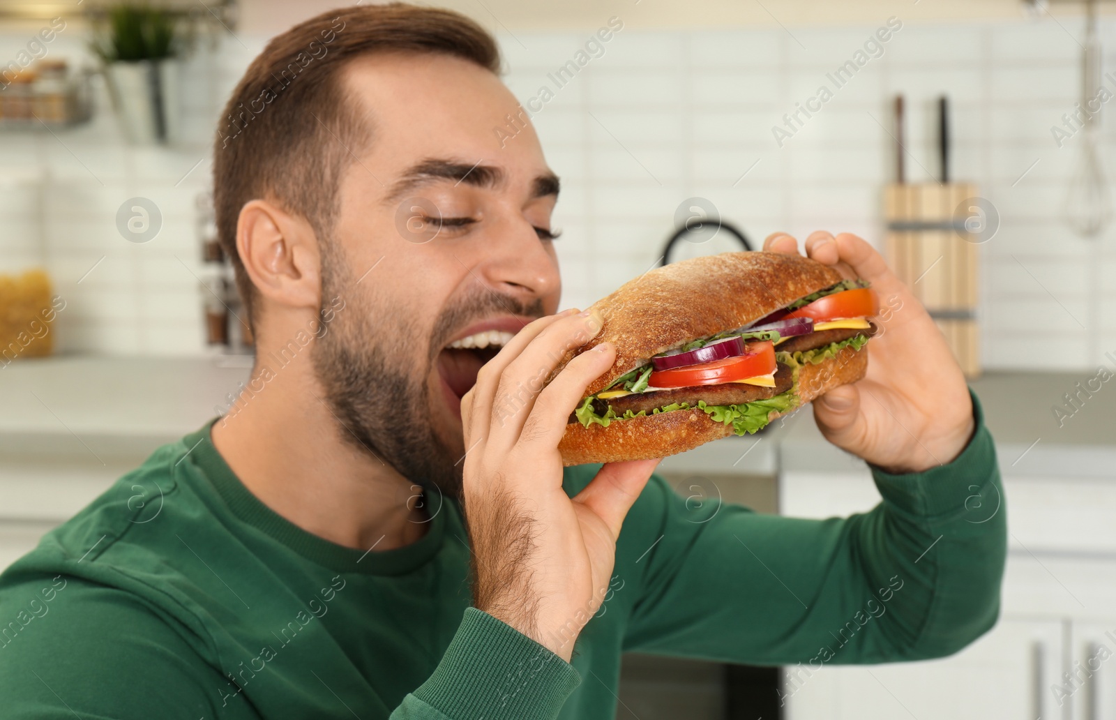 Photo of Young hungry man eating tasty sandwich in kitchen