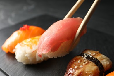 Taking delicious nigiri sushi with chopsticks from serving board on black table, closeup