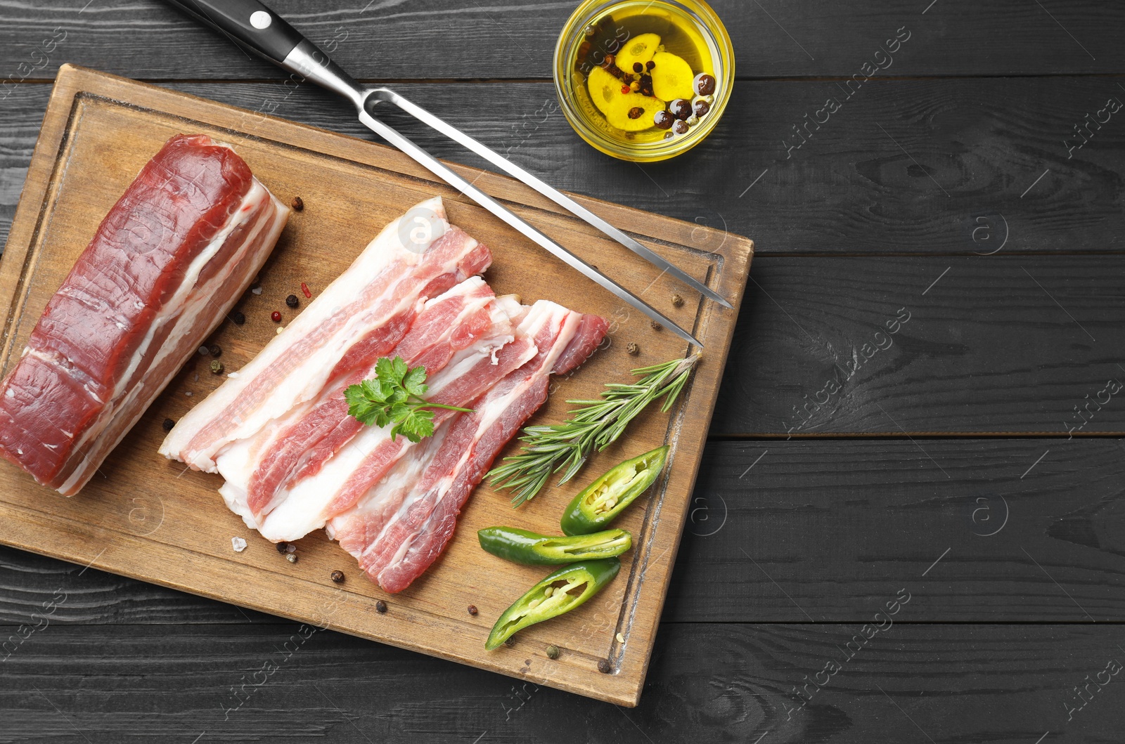 Photo of Pieces of raw pork belly, chili pepper, peppercorns, rosemary and parsley on black wooden table, flat lay