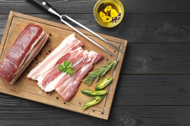 Pieces of raw pork belly, chili pepper, peppercorns, rosemary and parsley on black wooden table, flat lay