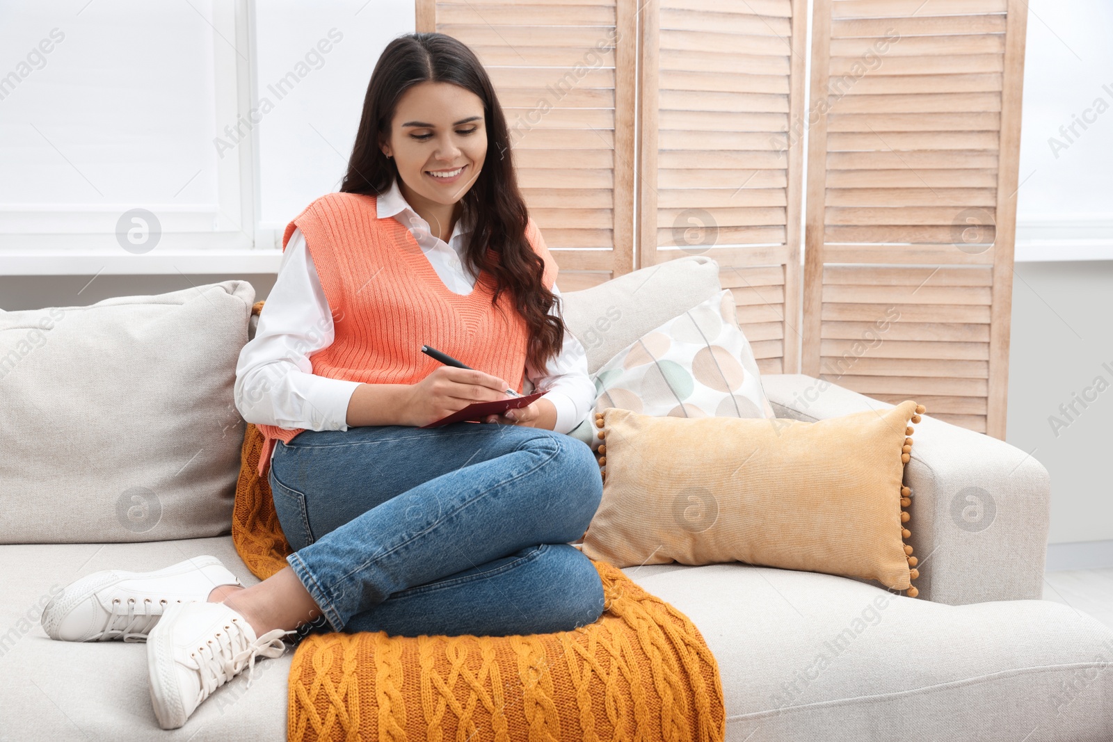 Photo of Young woman writing message in greeting card on sofa indoors