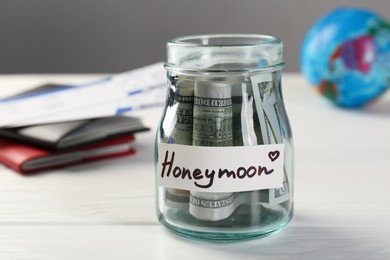 Glass jar with word Honeymoon and dollar banknotes on white wooden table, closeup