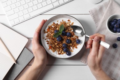 Photo of Woman eating tasty granola with blueberries at white wooden table with computer keyboard, top view