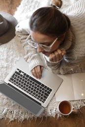 Photo of Woman with cup of hot beverage using laptop at home in winter evening, top view