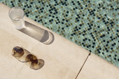 Photo of Stylish sunglasses and glass of water near outdoor swimming pool on sunny day, above view. Space for text