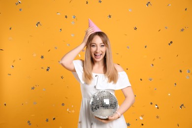 Happy young woman in party hat with disco ball and confetti on yellow background