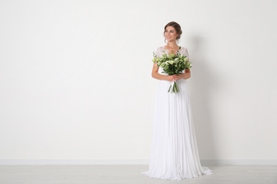 Young bride wearing wedding dress with beautiful bouquet near light wall. Space for text