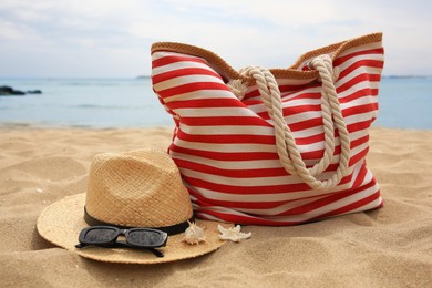 Stylish striped bag with straw hat, sunglasses, seashell and starfish on sandy beach near sea