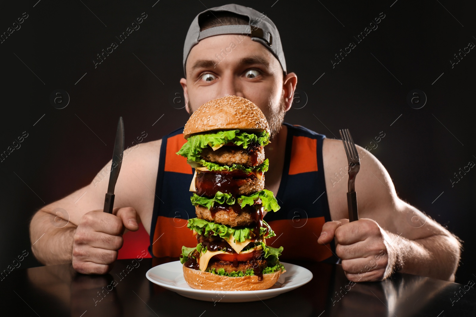 Photo of Young hungry man with cutlery eating huge burger on black background