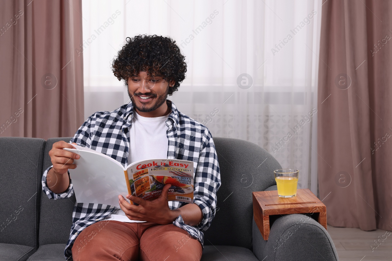 Photo of Happy man reading magazine at home. Glass of juice on wooden sofa armrest table