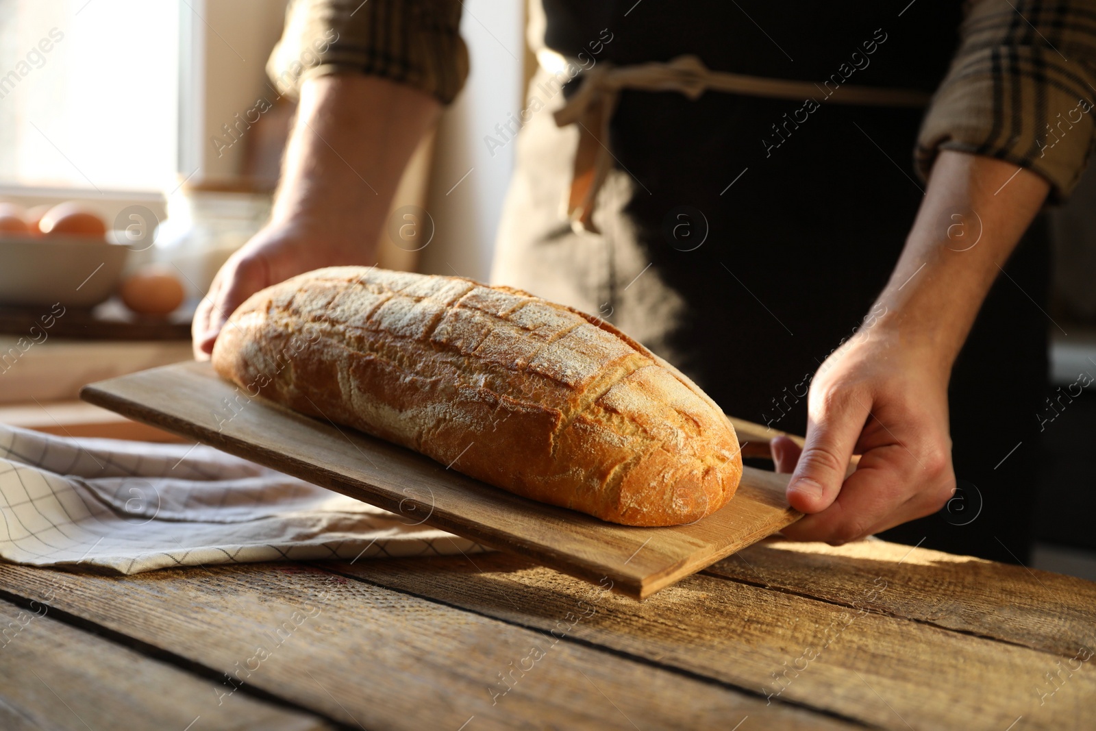 Photo of Man holding loaf of fresh bread at wooden table indoors, closeup