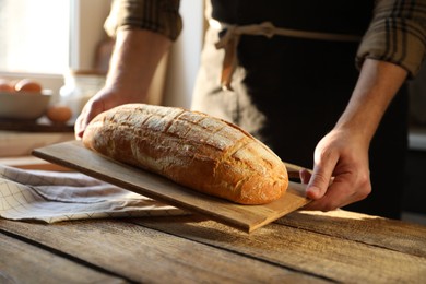 Photo of Man holding loaf of fresh bread at wooden table indoors, closeup